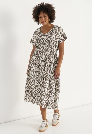 Kobieta ubrana w brązowo-białą, wzorzystą sukienkę midi z krótkimi rękawami i trampkami, stojąca na jednolitym białym tle.