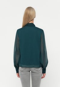 Τυρκουάζ μπλούζα με ημιδιάφανα μακριά μανίκια και κουμπιά στους καρπούς. Διαθέτει γιακά, ίσια στύλ και ελαφρύ, ρέον ύφασμα. Εμφανίζεται η οπίσθια όψη.