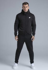 Musta vetoketjulla varustettu huppari ja siihen sopivat pehmeästä kankaasta valmistetut verkkarit. Rinnassa logo, resorit hihansuissa ja rento istuvuus.