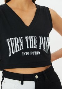 Cropped top nero con scollatura a V caratterizzato da testo grafico bianco "TRASFORMA IL DOLORE IN POTERE." Tessuto morbido e design senza maniche.