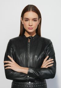 Czarna skórzana kurtka z zamkiem z przodu, wysokim kołnierzem, pikowanymi detalami na ramionach oraz srebrnymi ćwiekami wzdłuż dolnej krawędzi. Gładka tekstura.