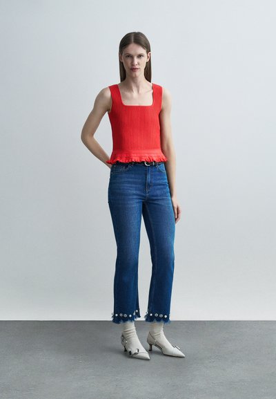 Γυναίκα που στέκεται φορώντας κόκκινο αμάνικο τοπ, μπλε κοντά τζιν με φλοράλ κρόσσια, λευκές κάλτσες και λευκά μυτερά παπούτσια με χαμηλό τακούνι.