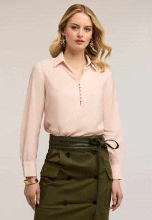 Femme aux cheveux blonds ondulés portant un chemisier rose pâle à manches longues, rentré dans une jupe taille haute couleur vert olive avec des boutons et une ceinture nouée.