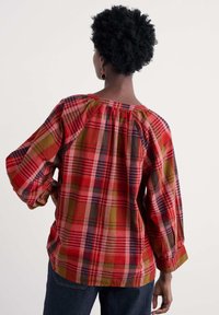 Czerwona i zielona koszula w kratę z długimi rękawami i marszczonym dekoltem. Materiał ma pomarszczoną teksturę i luźny krój.
