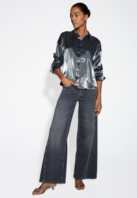 Donker metalen oversized shirt met een knoopsluiting aan de voorkant, gecombineerd met brede grijze jeans en zilveren sandaaltjes met bandjes. Glad, reflecterend textuur.