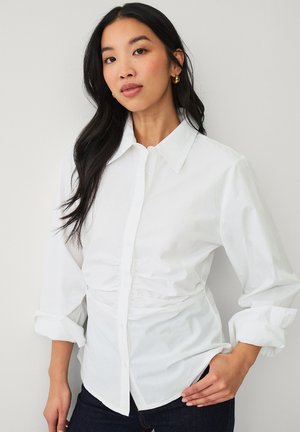 Kobieta z długimi ciemnymi włosami, nosząca złote kolczyki koła oraz białą koszulę zapinaną na guziki, z marszczonym pasem i podwiniętymi rękawami.