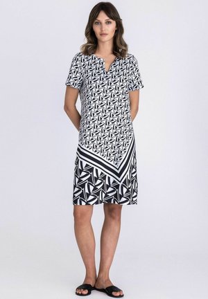 Femme portant une robe mi-longue à motifs géométriques noirs et blancs, à manches courtes et col en V, debout devant un fond clair uni.