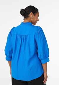 Hele sinine pluus lühikeste puffervarrukatega, voltidega kaunistatud õlad ja voltidega tagumine yoke. Kerge ja sileda tekstuuriga.