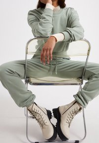 Sweat-shirt vert clair et joggings assortis, portés avec des bottines crème à lacets, dotées d'une semelle épaisse et de ferrures argentées, assis sur une chaise transparente.