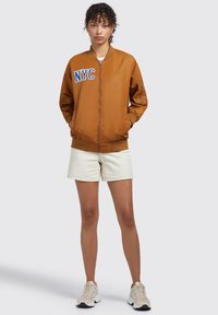 Giacca bomber marrone con ricamo "NYC" in blu, dotata di chiusura con zip, polsini a costine e tasca laterale; indossata sopra un outfit crema.