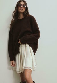Hnedý oversized sveter skombinovaný s bielou sukňou s krajkovým lemovaním. Textúra je mäkká; sukňa má plisovaný dizajn a krajkový okraj.