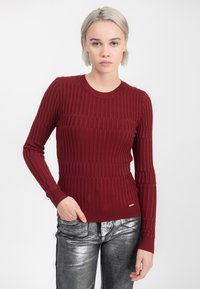 Vrouw met kort blond haar, gekleed in een gestructureerde bordeauxrode trui en metallic zilveren broek, staande tegen een effen witte achtergrond.