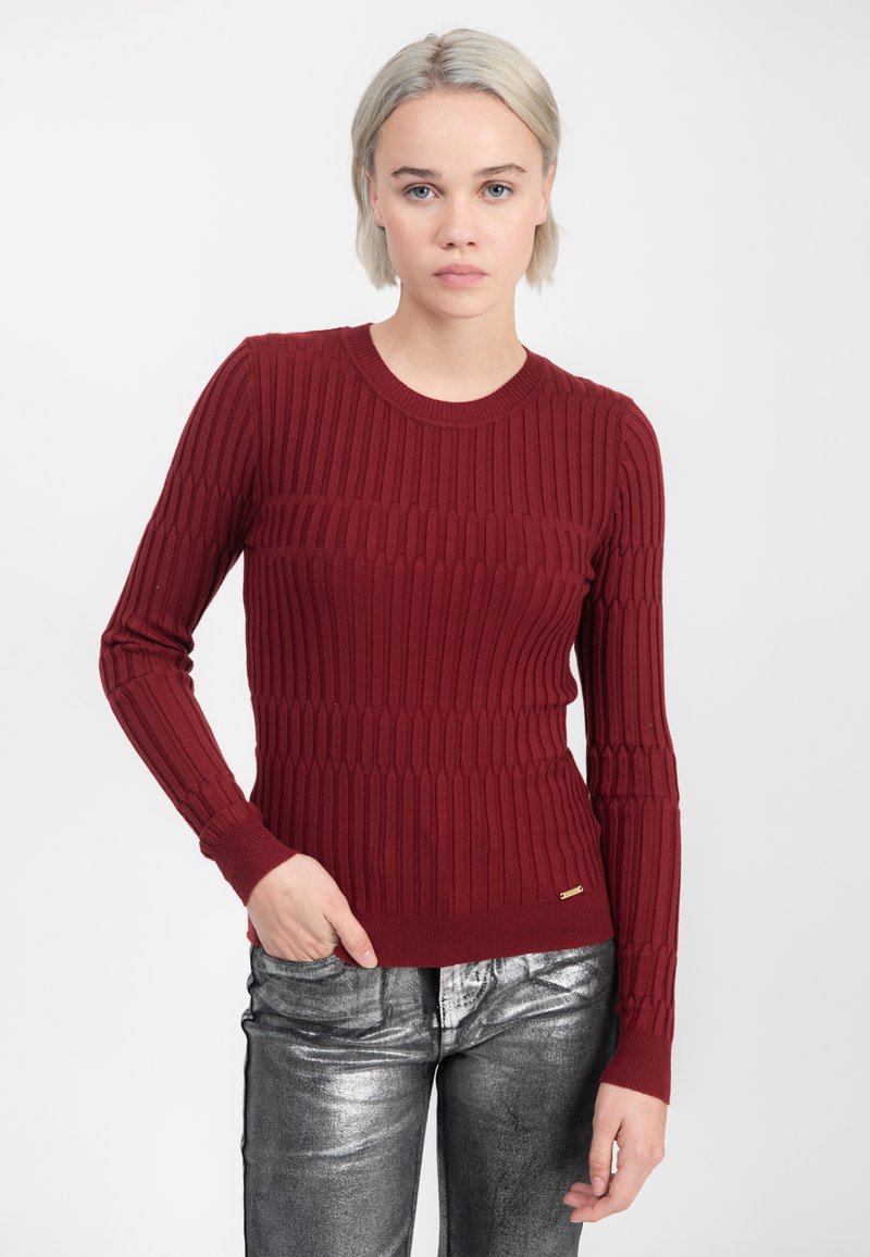 Vrouw met kort blond haar, gekleed in een gestructureerde bordeauxrode trui en metallic zilveren broek, staande tegen een effen witte achtergrond.