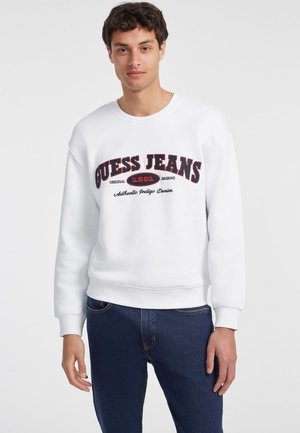 Felpa bianca con scollo rotondo, polsini e orlo a costine. Presenta una grafica "GUESS JEANS" in testo rosso e nero sulla parte anteriore.