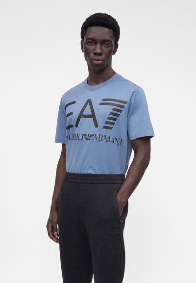 Νέος άνδρας με μπλε μπλουζάκι EA7 Giorgio Armani και μαύρο παντελόνι, με το ένα χέρι στην τσέπη, στέκεται μπροστά σε λευκό φόντο.