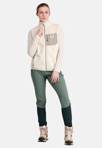 Fleecejacke in Creme mit einer grauen Brusttasche, kombiniert mit grünen Hosen und beigen Wanderschuhen, mit dunkelgrünen Akzenten an den Bündchen.