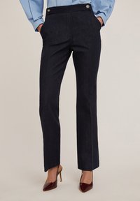 Marineblauwe broek met een getailleerde pasvorm, voorzien van zijzakken, gouden knopen op de tailleband en een gladde textuur. Draagt met bordeauxrode hakken.
