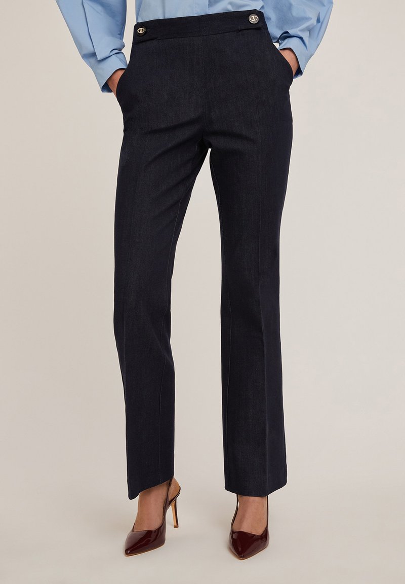 Marineblauwe broek met een getailleerde pasvorm, voorzien van zijzakken, gouden knopen op de tailleband en een gladde textuur. Draagt met bordeauxrode hakken.