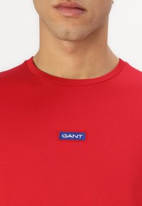 Piros pamut póló kerek nyakkal és rövid ujjakkal. Elöl egy hímzett, kék téglalap alakú címke található, rajta a "GANT" márkanevével.