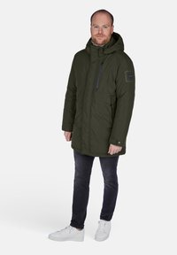 Olivgrön vadderad parkas med huva, dragkedjeförsett bröstfack, sidofickor och marinblå accenter. Bärs över mörka jeans och vita sneakers.