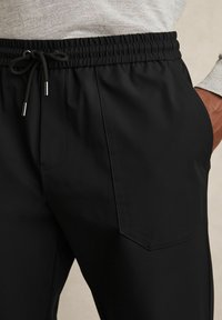 Black, lightweight trousers with an elastic waistband and drawstring. Features front pockets, smooth texture, and tailored fit.