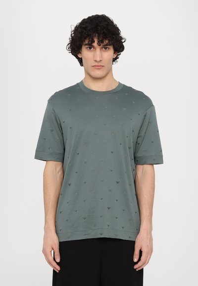 Maglietta di cotone verde con maniche corte, caratterizzata da un motivo sottile di piccole forme triangolari scure su tutto il tessuto.