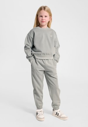 Junges Mädchen steht mit den Händen in den Taschen, trägt ein hellgraues Sweatshirt, passende Jogginghose und weiße Sneakers mit schwarzen Streifen.
