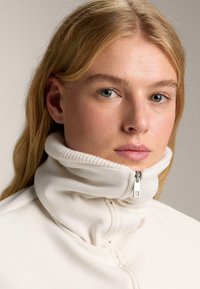 Biely kabát s vysokým golierom, so zapínaním na zips a rebrovaným golierom, charakterizovaný jemnou textúrou a hladkým povrchom. Pozadie v telovej farbe.