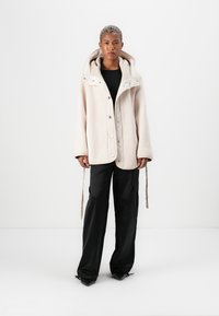 Cream-colored fleece jacket with a large hood, snap buttons, and two drawstrings. Worn over a black top and paired with black pants.