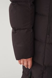 Donkerbruine puffer jack met gestikt textuur, met een hand zichtbaar die de mouw vasthoudt, geperste merkembleem op de mouw en een ritsluiting.