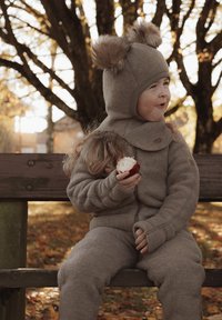 Grauer Fleece-Strampler mit Kapuze und Fellpompons, langen Ärmeln und Reißverschluss. Kind sitzt auf einer Bank, hält einen roten Apfel und ist von Herbstlaub umgeben.