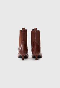Botas de tobillo de cuero marrón con tacones kitten, paneles laterales elásticos y tiradores, mostradas desde la parte trasera sobre un fondo blanco.