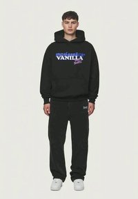 Črna kapuca z besedilom "Projection Vanilla Soft!" v pisanih črk, v kombinaciji s črnimi hlačami in belimi superge. Preprost, sproščen kroj.
