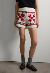 Pantalones cortos de punto en color crema con un patrón floral rojo, con detalles negros alrededor del dobladillo. Material texturizado, ajuste holgado, combinados con calcetines grises y zapatos negros.
