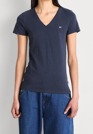 Kvinde iført en simpel mørkeblå T-shirt med V-hals og korte ærmer samt løstsiddende blå jeans, stående mod en lys grå baggrund.