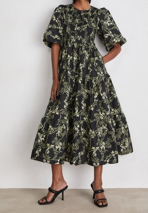 Robe midi noire avec motifs floraux verts, manches courtes bouffantes, taille cintrée, jupe à volants, portée avec des sandales noires à talons hauts.
