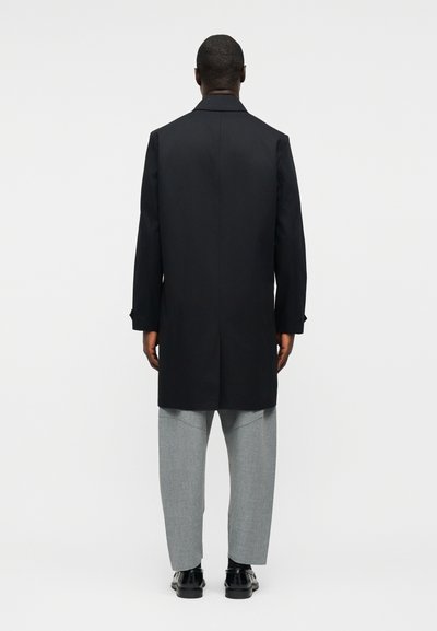 Άνδρας με μαύρο μακρύ παλτό, γκρι παντελόνι και μαύρα επίσημα παπούτσια, stoi με την πλάτη προς τα εμάς σε απλό λευκό φόντο.