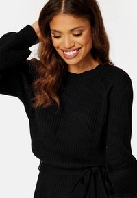 Pull en maille côtelée noir avec manches longues et col rond. Présente un détail de taille à nouer. Texture douce avec une silhouette ajustée.