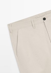 Pantalon beige sur mesure avec poches avant, passants pour ceinture, et fermeture à bouton noir sur fond blanc.