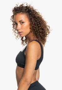 Top sportivo nero a coste con spalline spesse, finitura texturizzata e design aderente, mostrato in profilo laterale su uno sfondo bianco.