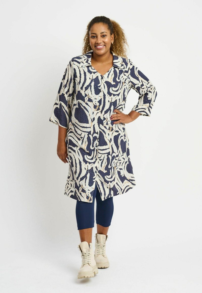 Long navy and white patterned shirt dress with a collar, drop shoulders, and a button-down front, paired with navy leggings and beige boots.