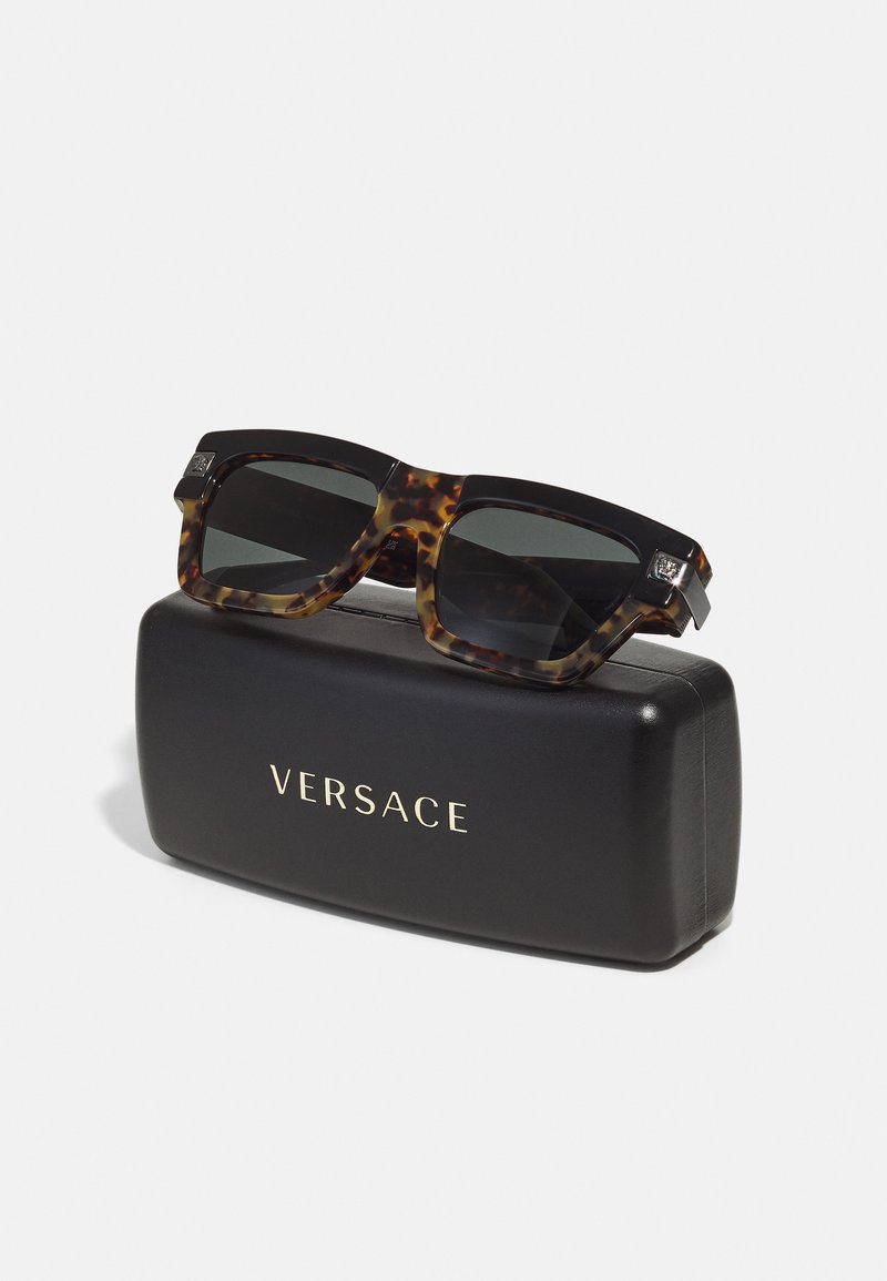 Solschar med sköldpaddsdesign, mörka linser, fyrkantiga bågar och silverdetaljer, vilande på ett svart Versace-fodral med guldtryck.