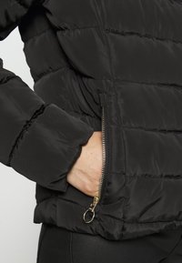 Crni puffer jakna s prugastim šavovima, patentnim zatvaračem, zlatnim dodacima i mekim, teksturiranim materijalom.