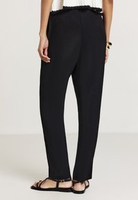 Femme portant un pantalon noir slim, des sandales plates noires à brides, un haut sans manches blanc et des bracelets en or, vue de dos.