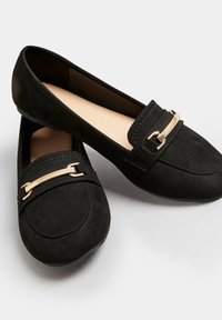 Mocassini neri in suede con suola piatta, caratterizzati da una barra decorativa dorata sulla parte superiore, cuciture a contrasto e una punta arrotondata.