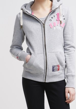 Persona vistiendo una sudadera con capucha gris claro con cremallera, con texto bordado en rosa y blanco y un bolsillo delantero, con las manos parcialmente en los bolsillos.