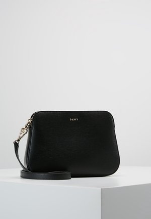 Petit sac bandoulière en cuir noir avec ferrures dorées et logo DKNY, posé sur un piédestal blanc sur un fond clair uni.