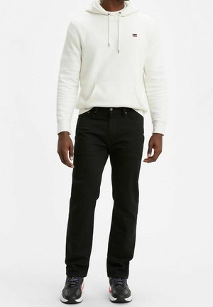 Man die een witte hoodie, zwarte jeans en zwarte sportschoenen draagt, staand tegen een effen witte achtergrond.