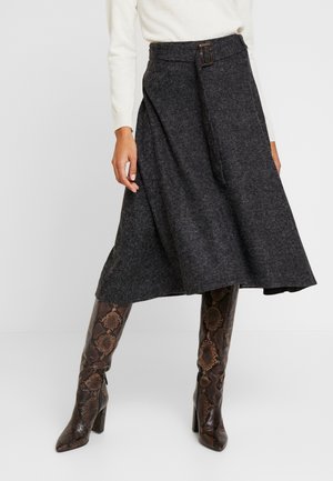 Vrouw draagt een donkergrijze wollen midi-rok met een gespsluiting, gecombineerd met kniehoge laarzen in een bruine slangenhuidprint.