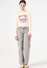 Sudadera con capucha crema y texto gráfico rojo, combinada con pantalones cargo grises de pierna ancha con bolsillos laterales. El modelo lleva zapatos beige.